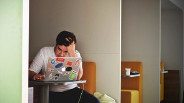 Person with a stressed expression sitting at a table, working on a laptop covered in stickers, illustrating anxiety and stress management in modern life.