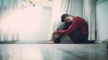 Young man sitting on the floor, hugging knees, appearing distressed in a bright room with sunlight filtering through sheer curtains, reflecting themes of anxiety and emotional struggles related to mental health.